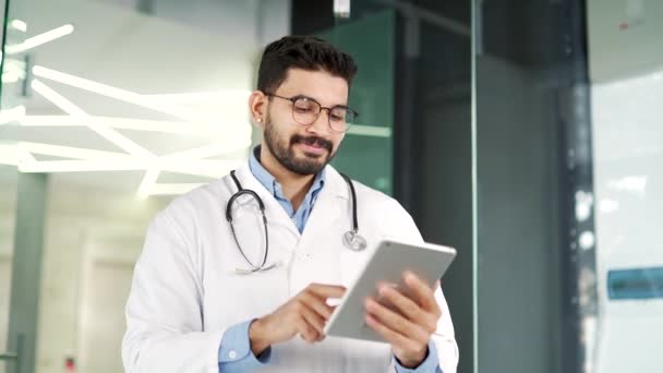 有信心的医生穿着白色外套站在医院诊所里用数码平板电脑 戴眼镜的大胡子医生与病人在网上聊天 在应用程序中工作或上网 — 图库视频影像