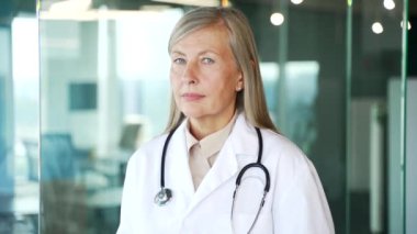 Hastane kliniğindeki kameraya bakan beyaz önlüklü yaşlı, gri saçlı kadın doktorun portresi. Ofiste poz veren kendinden emin, yaşlı bir doktor kadının vesikalığı. Kapat.