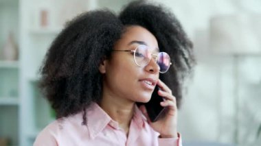 Mutlu Afro-Amerikan bayan öğrenci akıllı telefonuyla konuşuyor evdeki oturma odasında kanepede oturuyor. Gülümseyen siyah kadın bir meslektaşıyla konuşuyor ya da bir arkadaşıyla iletişim kuruyor. Kapat.