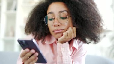 Depresyondaki genç Afro-Amerikan kadın evdeki oturma odasındaki kanepede otururken akıllı telefon kullanmaktan sıkıldı. Üzgün, yalnız siyahi kadın öğrenci esniyor, kötü bir ruh hali var, cep telefonunu karıştırıyor. Kapat.