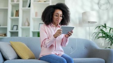 Genç, mutlu kulaklıklı Afro-Amerikan kadın evdeki oturma odasındaki kanepede otururken tablet üzerinde bilgisayar oyunu oynuyor. İlginç bir oyuna dalmış heyecanlı siyahi kadın öğrenci