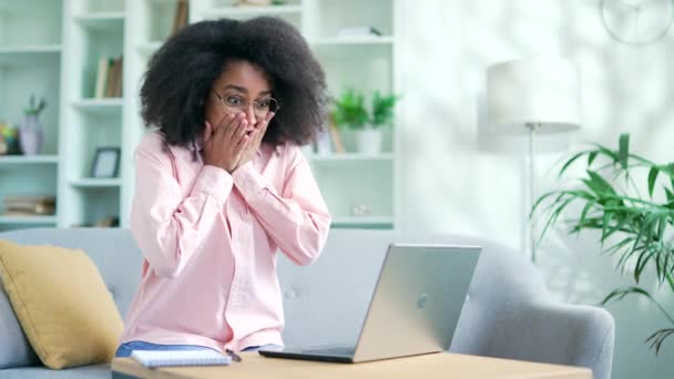 在家里的沙发上 坐在笔记本电脑上看好消息 让快乐的年轻非洲裔美国女性兴奋地庆祝成功 震惊的黑人女学生对计算机上的积极信息感到满意 — 图库视频影像