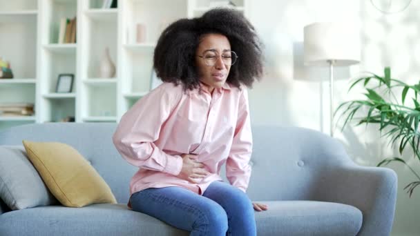 アフリカ系アメリカ人の不幸な若い女性は 自宅の部屋でソファーに座って腹痛に苦しんでいます 黒い女性は彼女の胃に手を置いています 彼女は中毒 スパムまたは月経を持っています — ストック動画