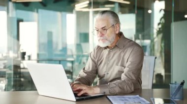 Kafası karışmış yaşlı, gri saçlı iş adamı iş yerinde dizüstü bilgisayar kullanmakta zorlanıyor. Kafası karışmış yaşlı erkeklerin bilgisayar uygulamalarında çalışırken karmaşık sorunları olur.