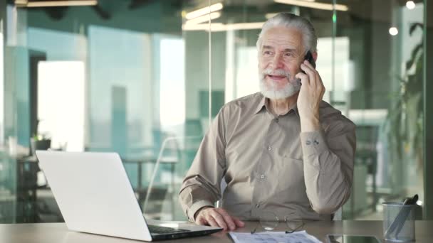 笑顔の幸せな上級灰色のひげ付きビジネスマンは 現代のオフィスで職場に座って電話で話しています 高齢者オーナーボスが会話を行い ビジネスパートナーやクライアントとコミュニケーションをとる — ストック動画