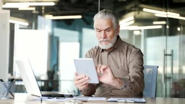 Ciddi düşünceli yaşlı, gri sakallı iş adamı iş yerinde dijital tablet kullanıyor. Odaklanmış yaşlı sahibi iletileri okur, istemciyle sohbet eder, uygulama bankacılığı yapar, internette gezinir