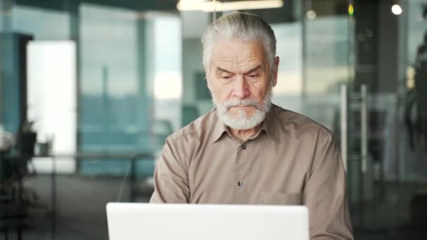 思慮深い上級グレーハイレッドのビジネスマンは ビジネスオフィスの職場に座っているノートパソコンで働いています 高齢の起業家は 問題の解決を考え コンピュータ上のプロジェクトで忙しい クローズアップ — ストック動画