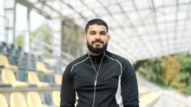 Bir stadyumda kameraya bakarak duran, kulaklıklı, gülümseyen sakallı bir sporcunun portresi. Eşofmanlı yakışıklı bir erkeğin kafasından. Yetişkin, mutlu sporcu antrenmanda. Kapat.