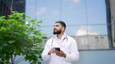 Beyaz önlüklü mutlu sakallı doktor hastane binasının önünde yürürken telefonu kullanıyor. Pozitif tıp doktoru bir hastayla internette sohbet ediyor ya da akıllı telefondan internette geziniyor.