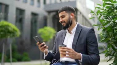 Mutlu iş adamı bir ofis binasının yakınındaki bir bankta oturan bir telefonu kullanıyor. Gülümseyen erkek elinde bir fincan kahve tutuyor, bir arkadaşıyla mesajlaşıyor, sohbet ediyor, sosyal medyada geziniyor.