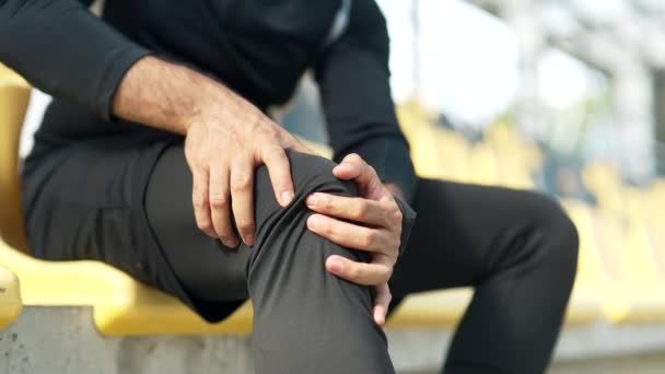 男性アスリートの手が膝をマッサージする スポーツマンは シティスタジアムに座っている筋肉痛を経験します 外でトレーニングしながら持続した外傷を軽減するために痛みを伴うスポットをこすります — ストック動画