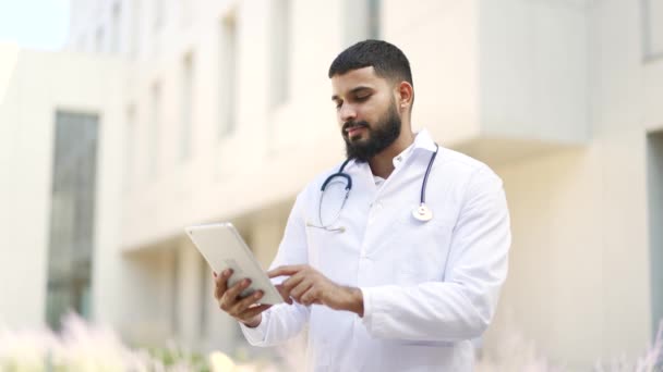 病院の建物の前に立っているデジタルタブレットを使用して白いコートの笑顔の医者 患者やブラウジングウェブでオンラインで口腔鏡検査を受けた肯定的な医療従事者医師 — ストック動画