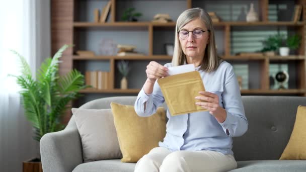 令人失望的高级灰白头发的女人坐在家里的沙发上阅读信件 带来了坏消息 一个令人不快的通知震惊于文件中的信息 使悲伤的退休老年妇女感到沮丧 — 图库视频影像