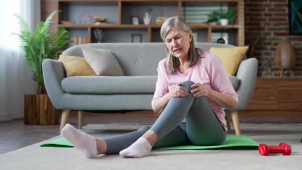在家中的客厅里 一位高个子灰发女子坐在垫子上进行体操锻炼 膝关节疼痛 疼痛严重 老年妇女按摩伸展创伤伤害 — 图库视频影像