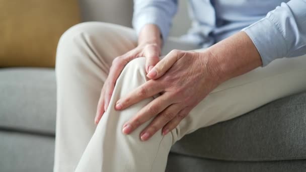 シニアの女性の手は 自宅のリビングルームでソファーに座って膝の筋肉をマッサージします 高齢の女性は痛みを伴う膝関節の痛みを患っており 歩くことができず 病気の痛み リウマチを患っています クローズアップ — ストック動画