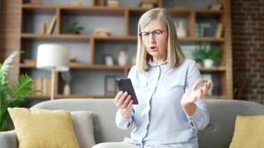 Kafası karışmış, gri saçlı, akıllı telefon kullanmakta zorlanan, evdeki oturma odasındaki kanepede oturan yaşlı bir kadın. Kafası karışmış yaşlı bir kadının telefondaki başvuru üzerinde çalışırken karmaşık bir sorunu var.