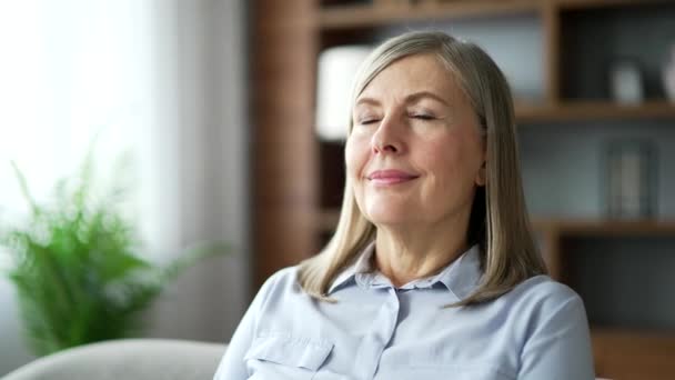 リビングに座っている間 目を閉じてリラックスしたシニア女性 自宅で快適なソファーで深呼吸をしている落ち着いた幸せな高齢女性の肖像画 瞑想コンセプト クローズアップ — ストック動画