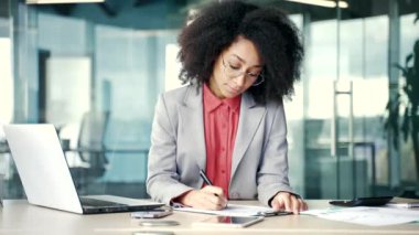 Meşgul genç Afro-Amerikalı iş kadını iş yerinde otururken belgeleri dolgu kalemle dolduruyor. Gözlüklü, evrak işleriyle uğraşan, vergi formu yazan siyahi kadın girişimci ya da finansör.