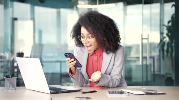 快乐而兴奋的年轻非洲裔美国女商人坐在办公室的工作场所 在电话里看到了好消息 黑人女人庆祝成功 为胜利而高兴 摆出姿势是的 — 图库视频影像