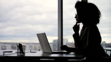 Siluet. Düşünceli, odaklanmış, genç, Afro-Amerikan bayan çalışan dizüstü bilgisayarda çalışıyor ve ofisteki iş yerinde problem çözmeyi düşünüyor. Ciddi siyahi kadın bir projeyle meşgul.