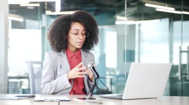 Genç Afrikalı Amerikalı iş kadını elinde mikrofonla ofiste oturan bir dizüstü bilgisayarla podcast yapıyor. Kendine güvenen bayan antrenör uzaktan eğitim veriyor, video aracılığıyla iletişim kuruyor.
