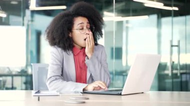 Çok çalışan Afro-Amerikan iş kadını yorgun hissediyor ve iş yerinde çalışırken dizüstü bilgisayarda uyumak istiyor. Uykulu yorgun siyah kadın esniyor, gözlerini kapıyor. İşte aşırı yüklenme