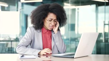 İş yerinde dizüstü bilgisayarla çalışırken baş ağrısı çeken yorgun Afrikalı Amerikalı iş kadını. Üzgün siyah kadın acı hissediyor, elleriyle şakaklarına masaj yapıyor.