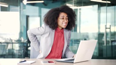 Dizüstü bilgisayarla çalışırken sırt ağrısı çeken Afrikalı genç bir iş kadını ofiste oturuyor. Üzgün siyah kadın elini beline koyup ağrıyan kaslarına masaj yapıyor.