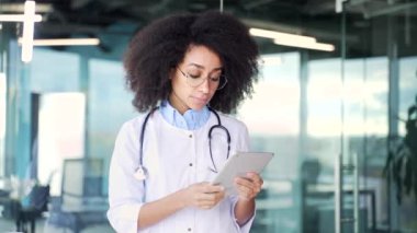 Beyaz önlüklü genç Afrikalı Amerikalı kadın doktor modern hastane kliniğinde dijital tablet kullanıyor. Tıp doktoru ofiste dururken bir hastayla sohbet ediyor. Kapat.