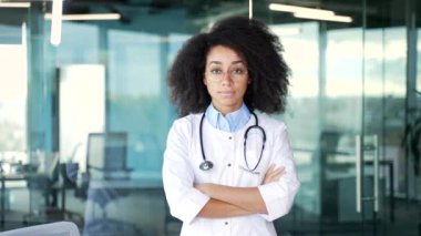 Modern hastane kliniğinde kameraya bakan beyaz önlüklü, ciddi, Afro-Amerikan kadın doktor portresi. Siyahi kadın doktor ofiste çapraz kollarla poz veriyor.