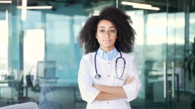 Hastane kliniğinde kameraya bakan beyaz önlüklü mutlu, Afro-Amerikan kadın doktor portresi. Gülümseyen siyahi kadın tıp doktoru ofiste kollarını kavuşturarak poz veriyor.