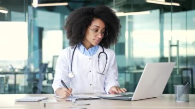 Beyaz önlüklü genç Afro-Amerikan kadın doktor hastane kliniğindeki iş yerinde dizüstü bilgisayara bakarak reçete yazıyor. Siyahi kadın tıp doktoru ofiste çalışıyor.