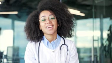 Webcam görüntüsü. Gülümseyen Afro-Amerikan kadın doktor hastane kliniğindeki kameraya bakarak konuşuyor. Mutlu bir hekim ofiste otururken hastasına danışıyor. Kapat.