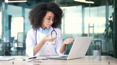 Mutlu Afro-Amerikan kadın doktor hastane kliniğinde dizüstü bilgisayarla konuşuyor. Gülümseyen genç tıp doktoru ofiste otururken hastasıyla uzaktan konsültasyon yapıyor.