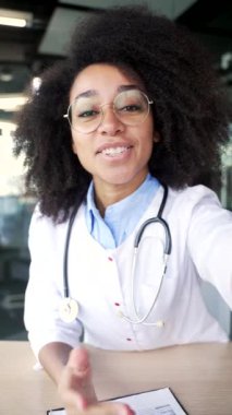 POV dikey video. Mutlu Afro-Amerikan kadın doktor bir hastane kliniğinde görüntülü konuşma yapıyor. Gülümseyen genç tıp doktoru hastasıyla kameraya bakarak sohbet ediyor.