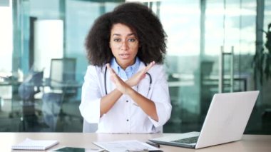 Genç Afro-Amerikan kadın doktor kollarını çapraz gösteriyor, X işareti, başını sallıyor hastane kliniğinde oturmuyor. Tıp doktoru bir yasaklama, inkar, anlaşmazlık jesti gösteriyor.