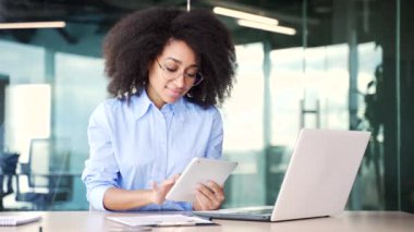 Genç Afro-Amerikan bayan çalışanı iş yerindeki iş yerinde dijital tablet kullanıyor. Gülümseyen siyah kadın bir mesaj yazıyor, bir müşteriyle sohbet ediyor ya da internette geziniyor.