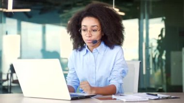 Kablosuz kulaklıklı Afro-Amerikan kadın müşteri temsilcisi ofiste otururken bilgisayarla video görüşmesi yapıyor. Çağrı merkezinin temsilcisi müşteriye tavsiyelerde bulunup soruları cevaplıyor.