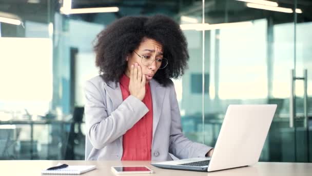 アフリカ系アメリカ人の若い実業家は オフィスの職場でノートパソコンの仕事に苦しんでいる 黒人女性は彼女の頬に彼女の手を保持しています 痛みの場所をマッサージ 歯科医に会う必要があります — ストック動画