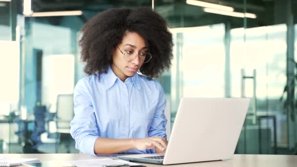 年轻的非洲裔美国女雇员坐在办公室工作时在笔记本电脑上打字 戴眼镜的黑人妇女从事计算机应用工作 在网上聊天或忙于某个项目 — 图库视频影像