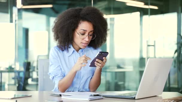 年轻的非洲裔美国女雇员坐在办公室工作时 在电话上看到坏消息时 感到很震惊 黑人妇女对信息 收到的通知或在网上看到的东西感到失望 — 图库视频影像