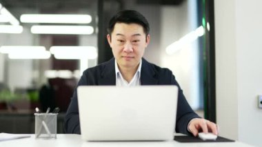 Memnun Asyalı işadamı iş yerinde otururken dizüstü bilgisayarından harika haberler aldı. Resmi takım elbiseli olgun bir adamın gülümsemesi bilgisayardan olumlu bir mesaj okuyor.