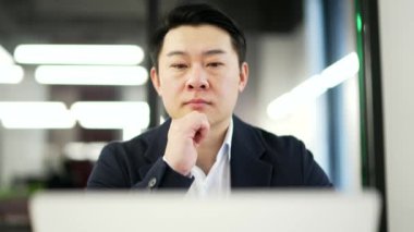 Düşünceli ciddi Asyalı işadamı iş yerindeki bilgisayarını kullanıyor. Resmi bir takım elbise sohbetinde odaklanmış bir adam, bilgisayar uygulamasında çalışıyor ya da başlangıç projesini düşünüyor. Kapat.