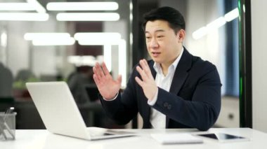 Kendine güvenen Asyalı iş adamı ofisteki bir iş yerinde dizüstü bilgisayarla konuşuyor. Resmi takım elbiseli bir adamın iş görüşmesi var, uzaktan konuşuyor, online bir konferansta iletişim kuruyor.