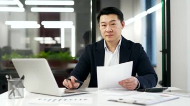 İş yerindeki çalışma masasında oturan resmi takım elbiseli mutlu bir Asyalı muhasebecinin portresi. Gülümseyen olgun bir finansörün kağıt işleriyle uğraşırken, dizüstü bilgisayarda çalışırken ve kameraya bakarken.