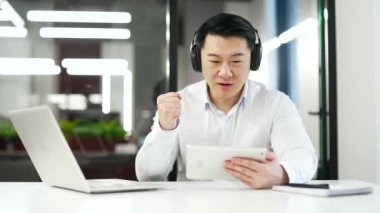 Kablosuz kulaklıklı heyecanlı Asyalı adam iş yerinde çalışırken dijital tablette spor müsabakaları izliyor. Mutlu olgun işçi duygusal olarak favori takım için tezahürat yapıyor.
