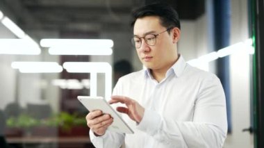 Ciddi Asyalı işadamı iş yerinde dikilirken dijital tablet kullanıyor. Gözlüklü adam mesajları okur veya yazar, istemci, bankacılık, e-posta kontrolü veya internette gezinme