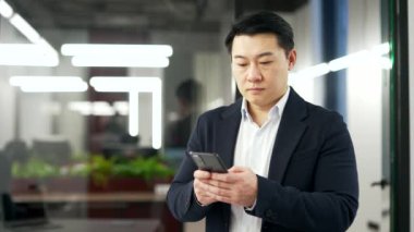 Ciddi Asyalı işadamı iş yerinde dururken telefonu kullanıyor. Resmi takım elbisedeki odaklanmış adam mesajları okur veya yazar, istemciyle konuşur, bankacılık veya akıllı telefondan internette gezinir