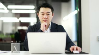 Düşünceli ciddi Asyalı işadamı iş yerindeki bilgisayarını kullanıyor. Resmi bir takım elbise sohbetinde odaklanmış bir adam, bilgisayar uygulamasında çalışıyor veya başlangıç projesini düşünüyor