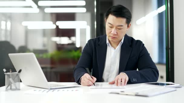 Busy Asianのビジネスマンは 職場に座っているラップトップコンピュータを使用して書類を作成しています 正式なスーツの仕事で自信を持って成熟した男は文書を記入し 執筆し ビジネスオフィスで税金のリターンを作ります — ストック動画